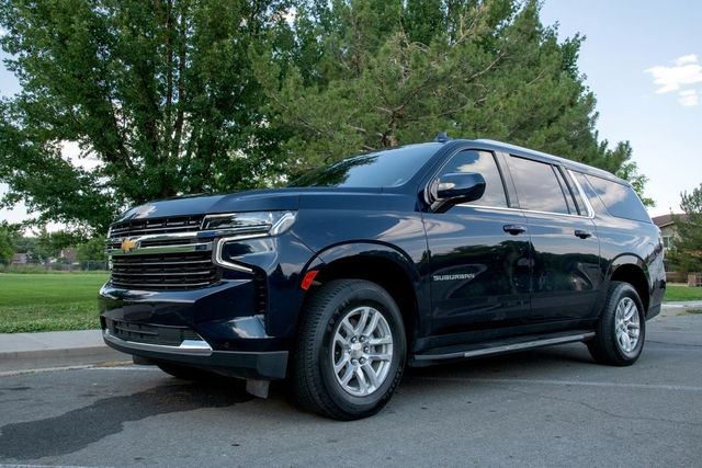 A dark blue Chevrolet Suburban parked on an asphalt road with green trees and a grassy field in the background.