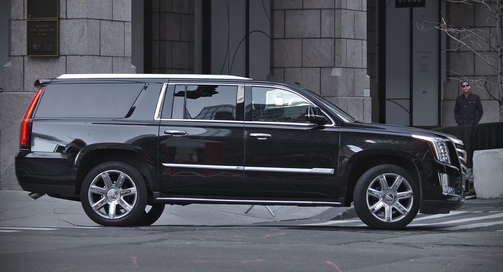 A black Cadillac Escalade ESV drives past a stone building on a city street.