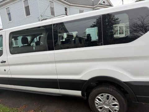 A side view of a white commercial passenger van parked on an asphalt street in front of a blue house.
