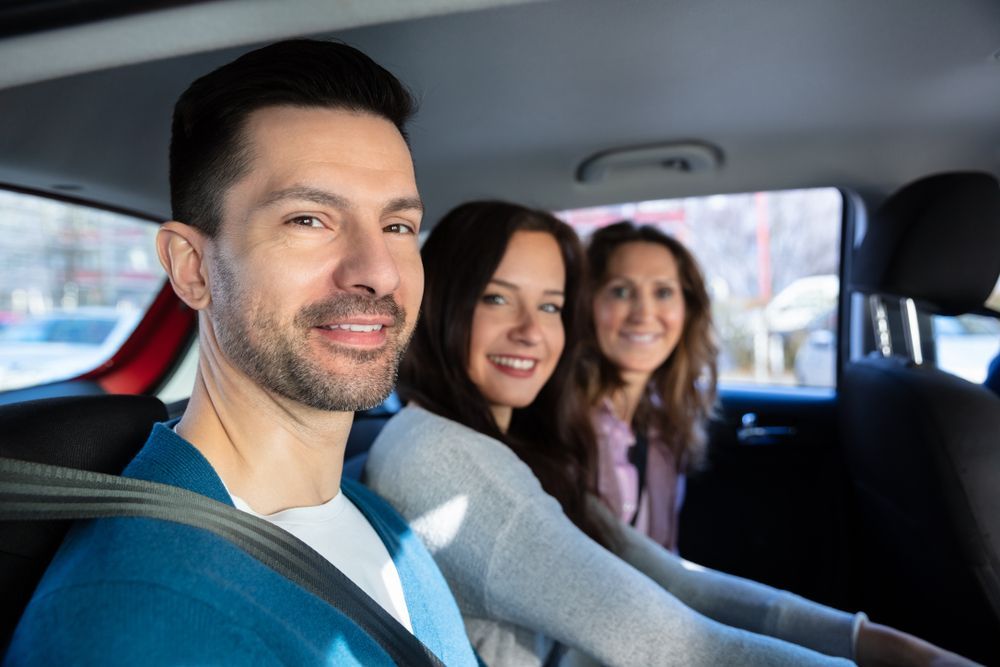 Three people smiling while sitting in the back seat of a car, wearing seatbelts.