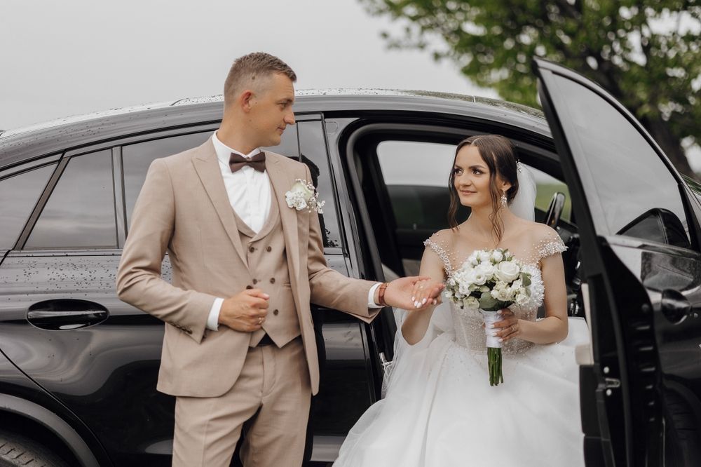 A groom in a beige suit reaches out his hand to help a bride in a white wedding gown step out of a black car.