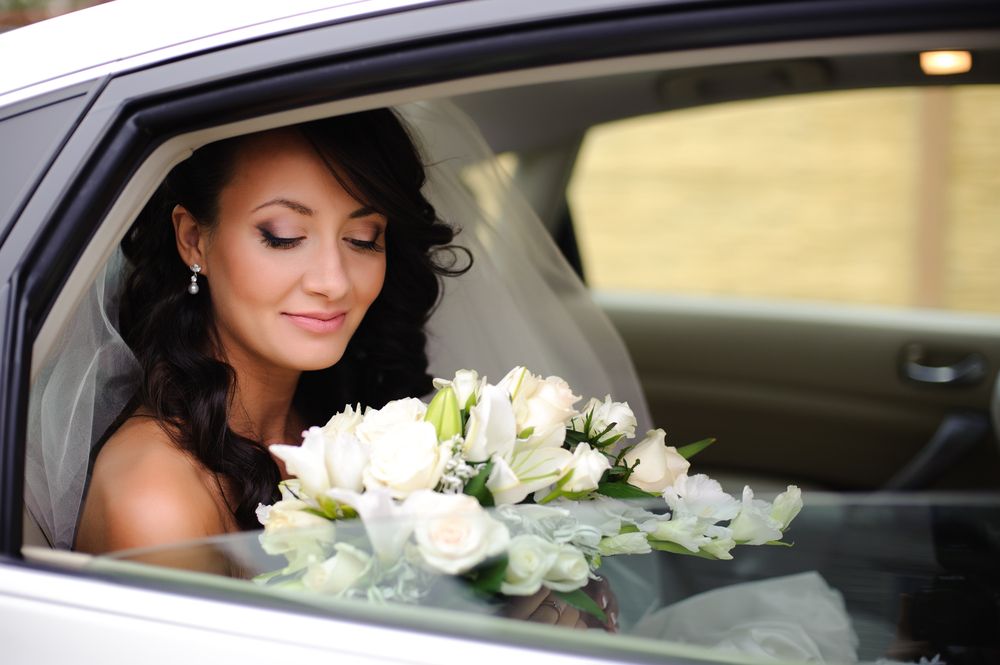 A bride with dark, wavy hair and a veil sits in a car, looking down at a bouquet of white roses.