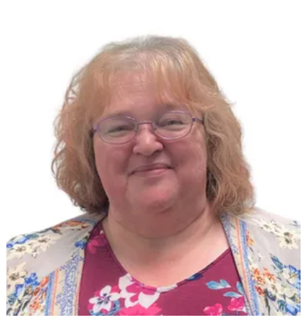 A smiling person with shoulder-length wavy hair and glasses, wearing a floral-patterned shirt and cardigan.