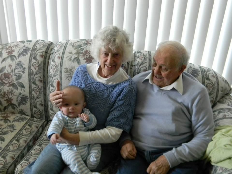 An older couple and a baby sit together on a patterned sofa in front of vertical blinds.