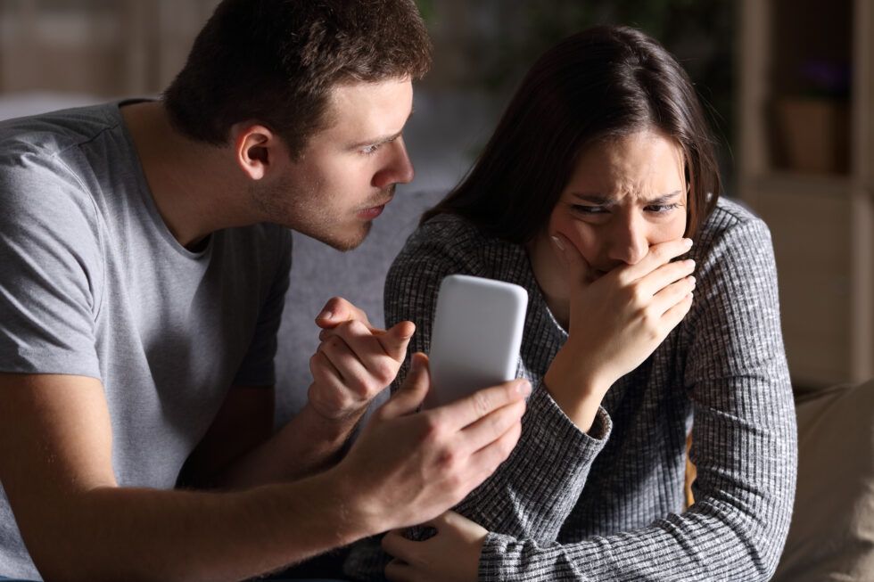 A person shows a phone to another person who looks distressed and covers their mouth with their hand.