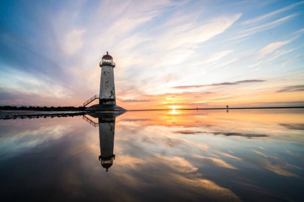 A lighthouse stands on a beach at sunset, its reflection clearly visible in the wet sand below.