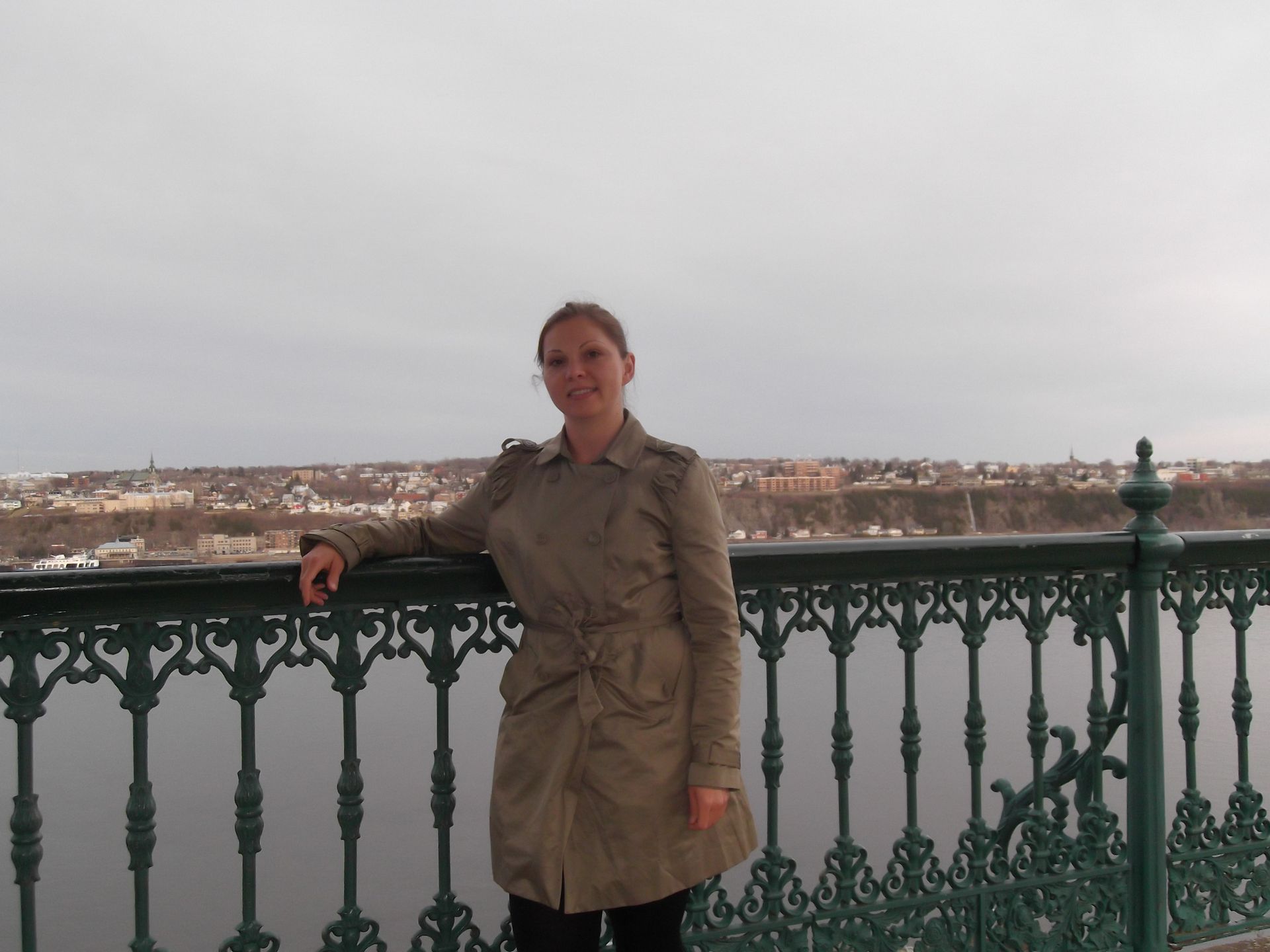 Woman in tan coat leans on ornate railing, overlooking cityscape and water on a cloudy day.