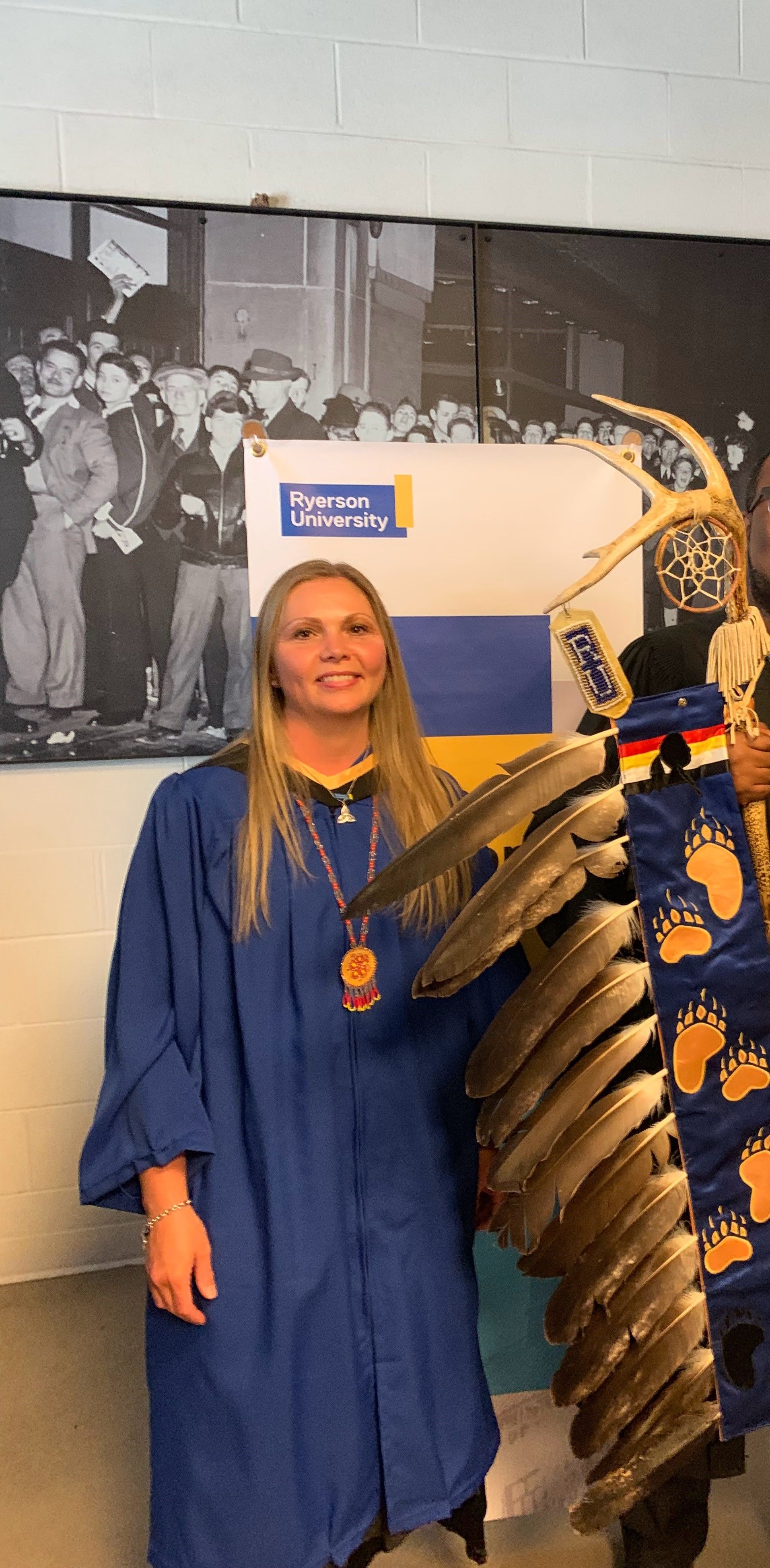 Woman in graduation gown with ceremonial feathers and Native American clothing. Blue and gold logo in the background.
