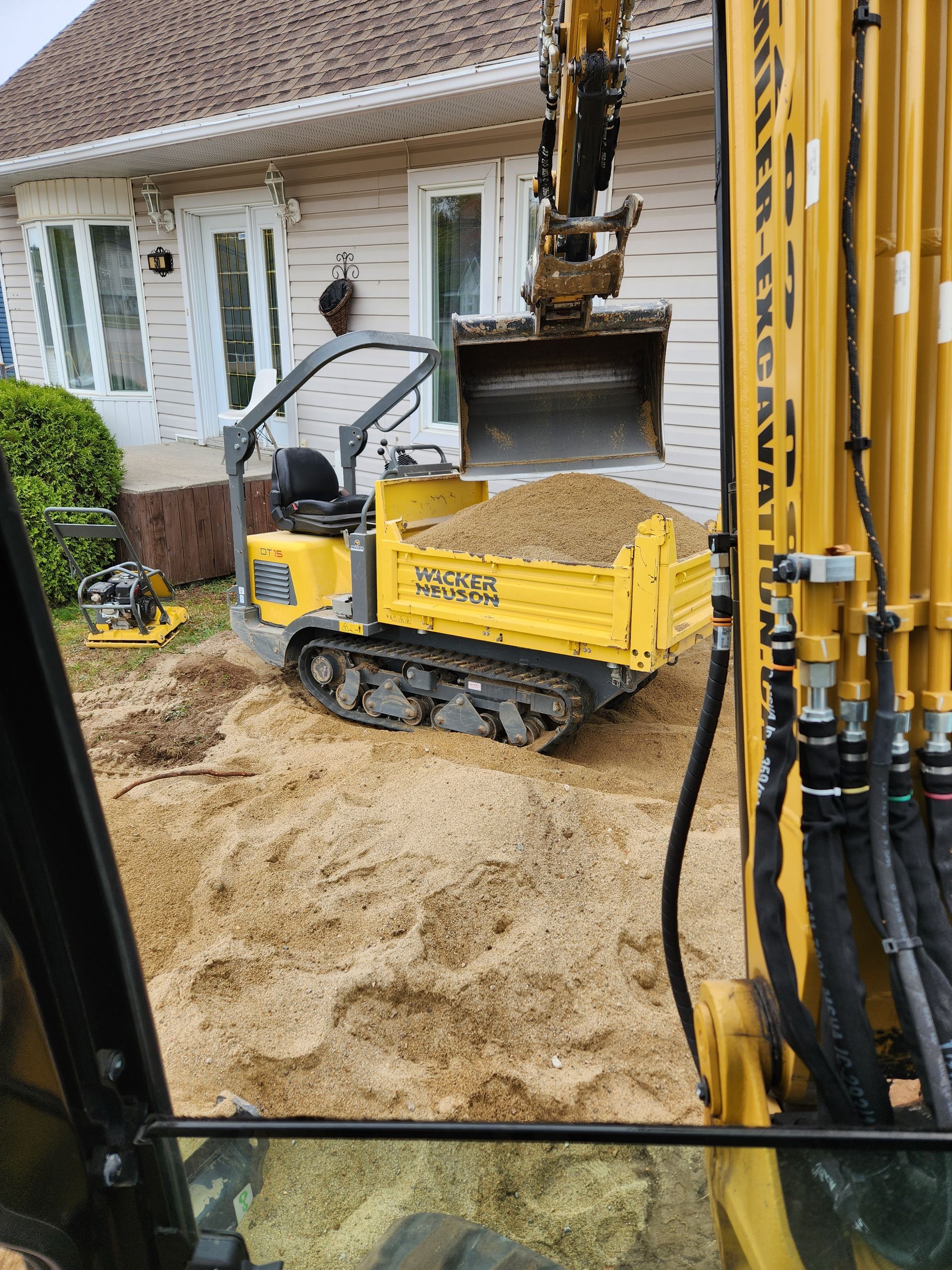 Une excavatrice jaune charge de la terre dans une benne à ordures devant une maison.