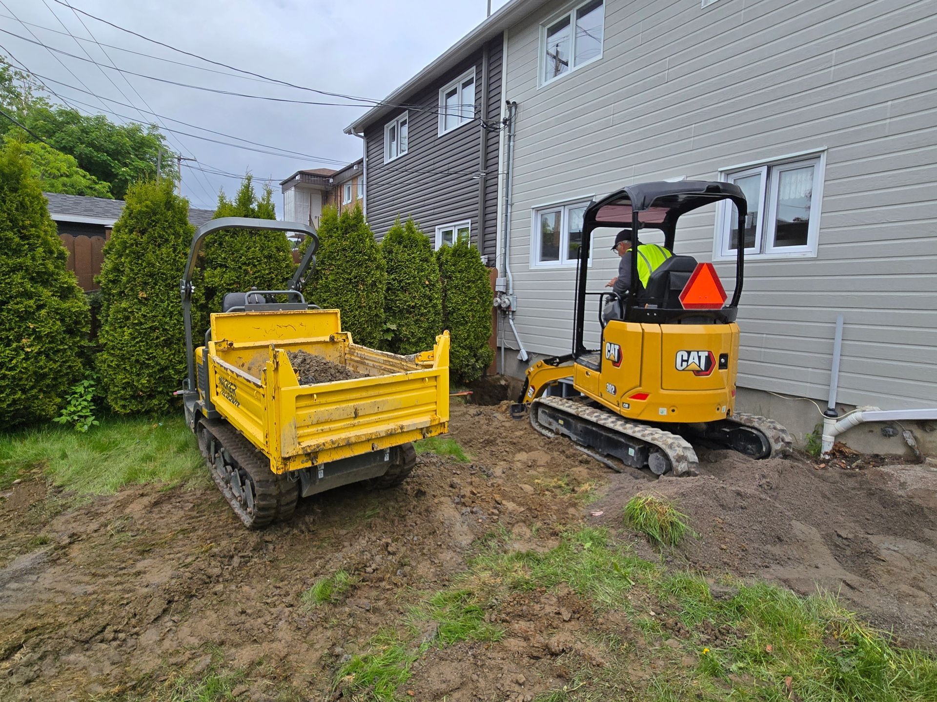 Un camion-benne jaune et une excavatrice jaune sont garés devant une maison.