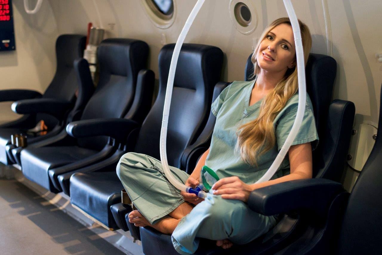 A woman in scrubs is sitting in a row of seats on an airplane. Hyperbaric Oxygen Therapy