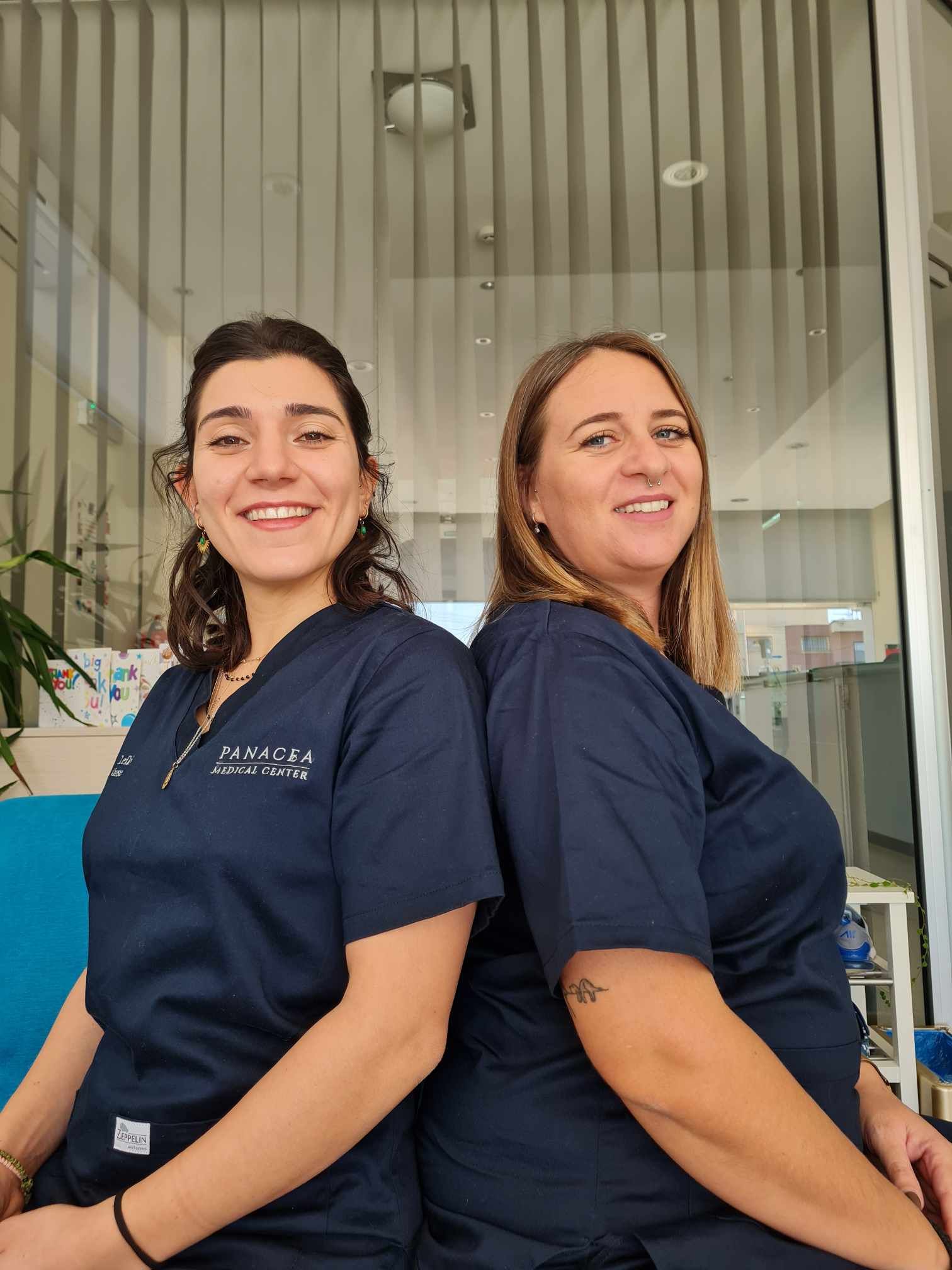 Two women in scrubs are sitting back to back and smiling.