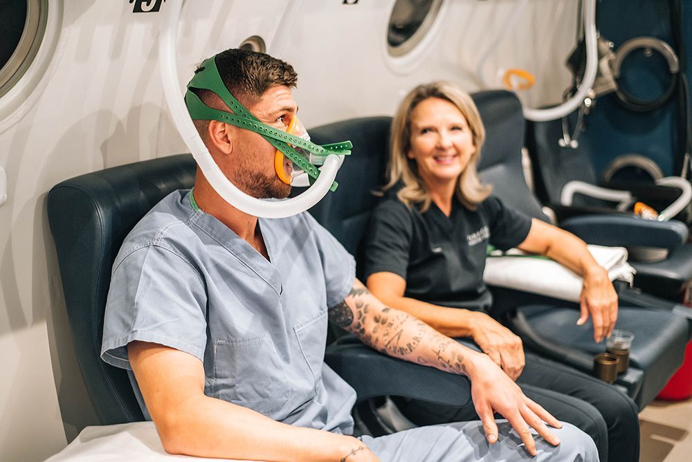 A man wearing an oxygen mask is sitting in a chair next to a woman. in a Hyperbaric Oxygen Therapy.