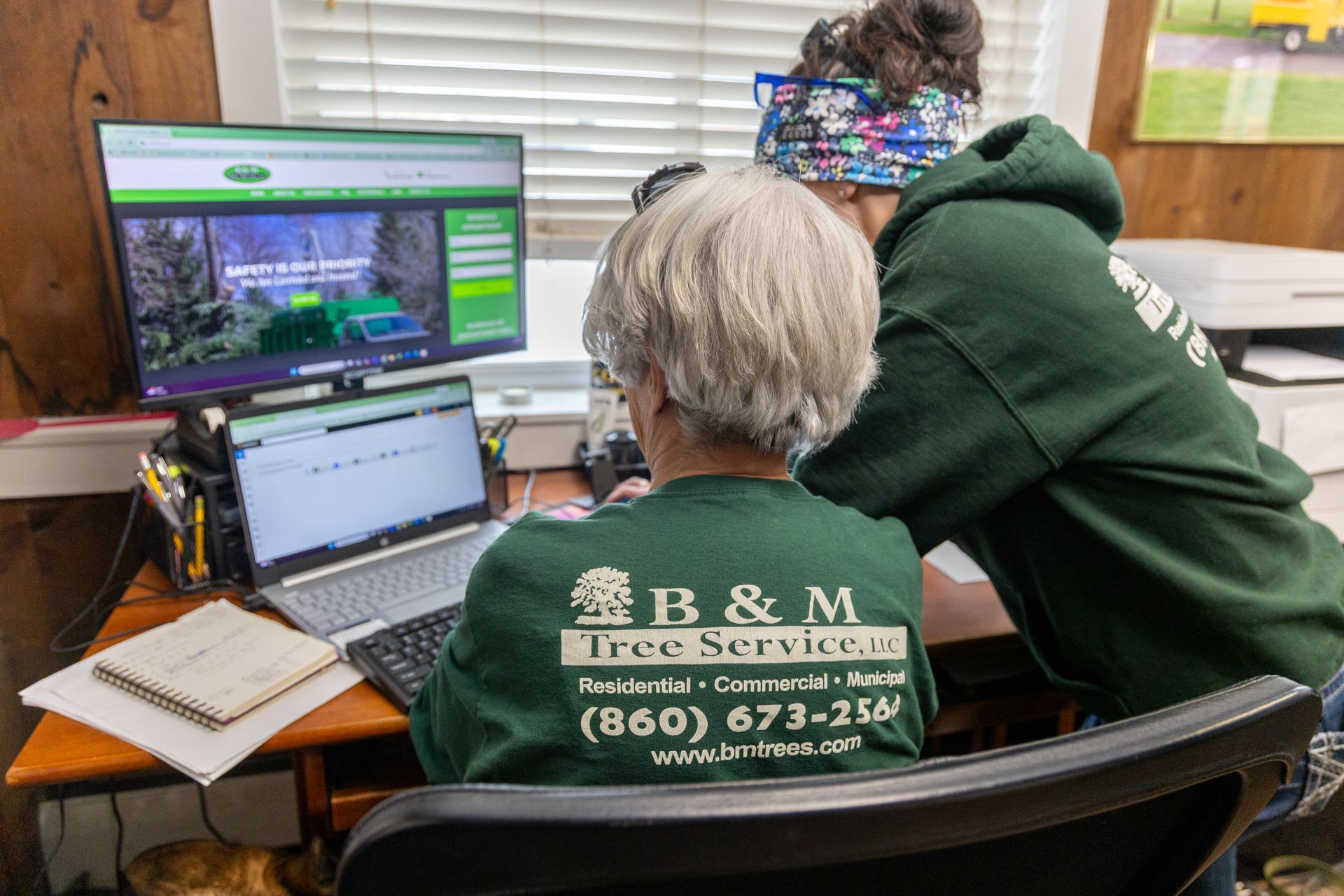 Two people wearing B&M Tree Service green shirts work together at desks with a computer monitor and laptop.