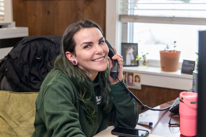 A person with green-tinted hair wearing a green sweater smiles while talking on a landline phone at an office desk.
