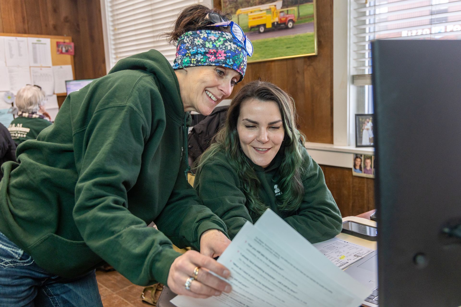 Two people wearing green hoodies collaborate while looking at a document in an office setting.