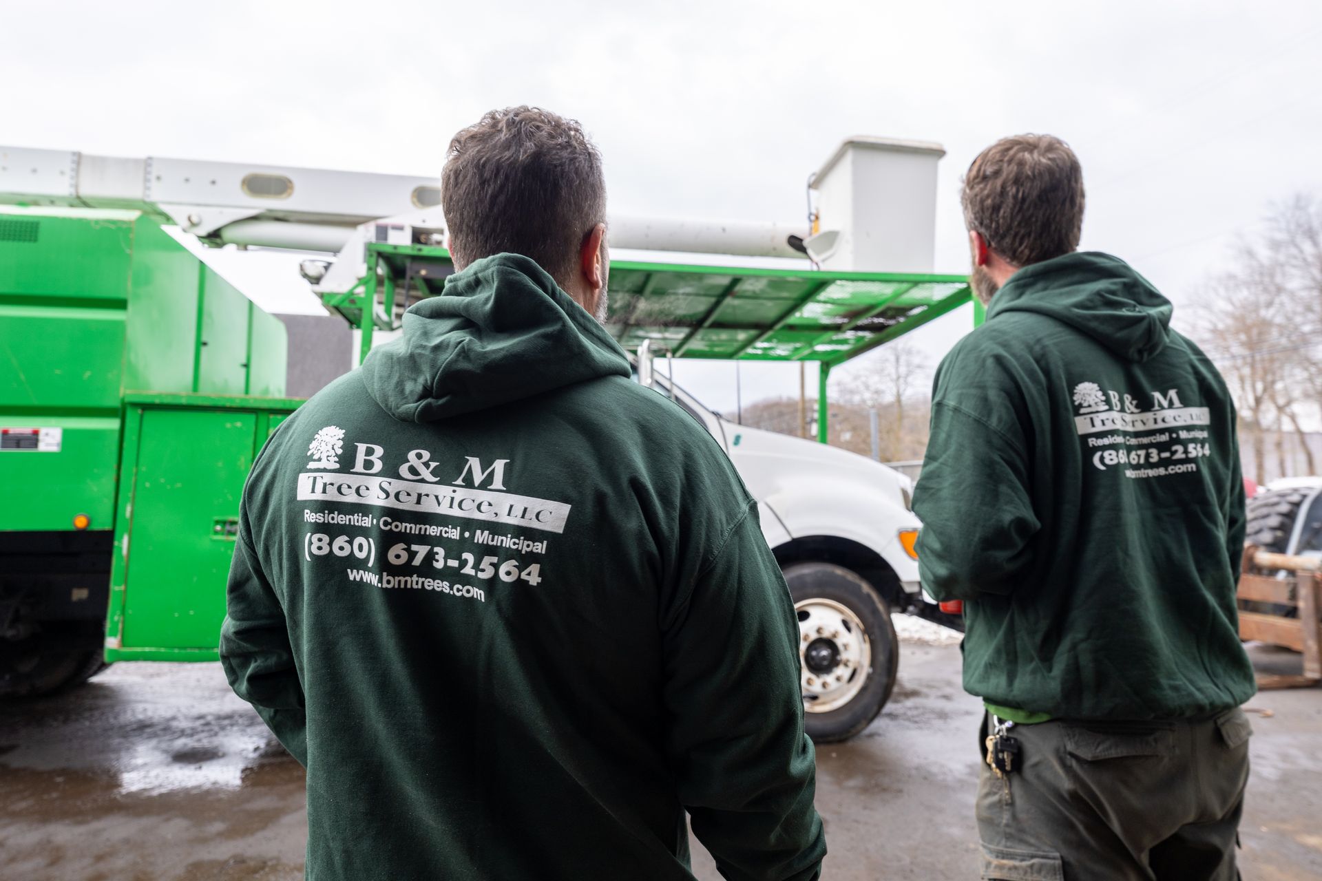 Two people in dark green B&M Tree Services hoodies stand outdoors facing a green utility truck with a crane.