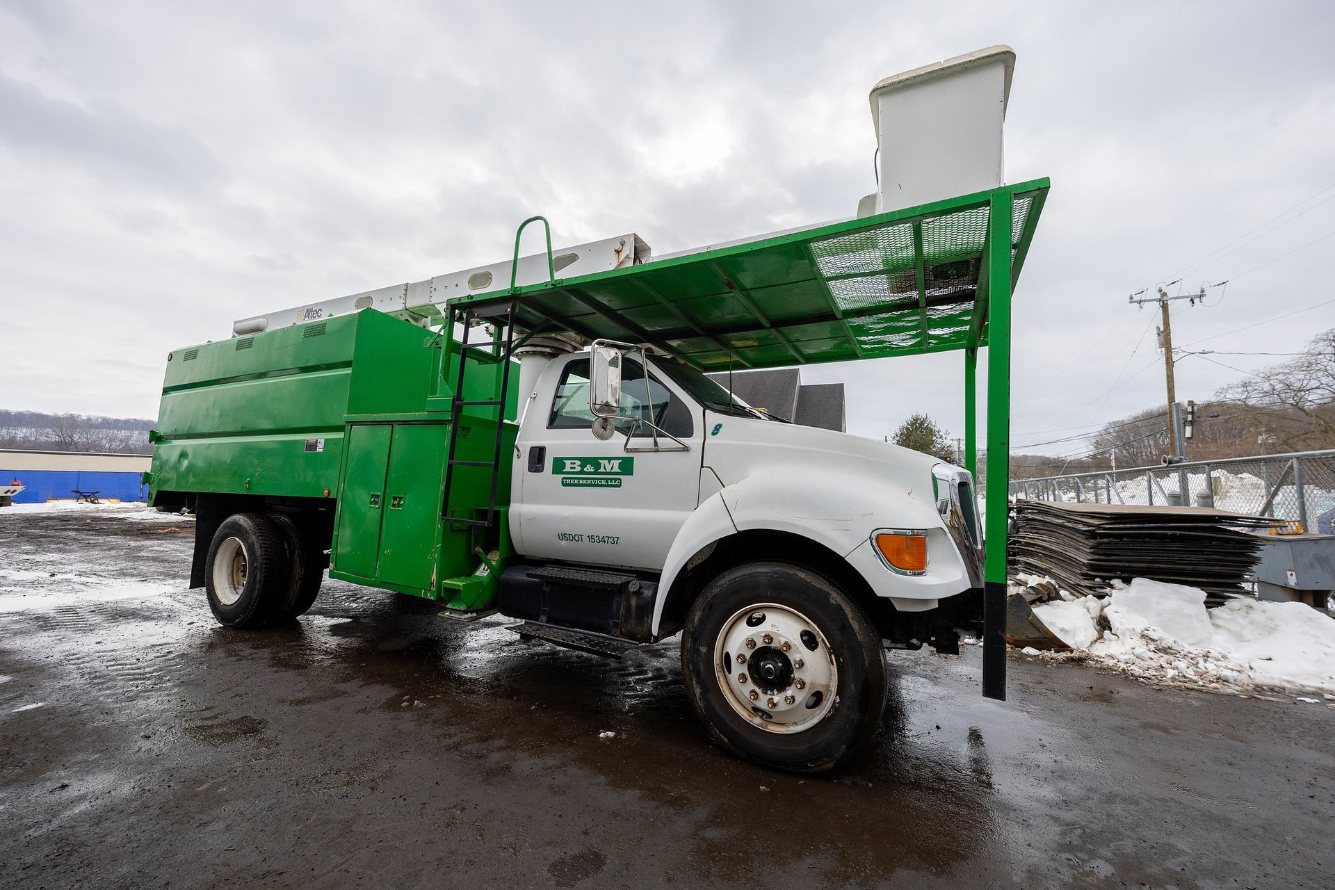 A green and white bucket truck parked on a muddy, snowy lot, featuring an extended frame over the cab.