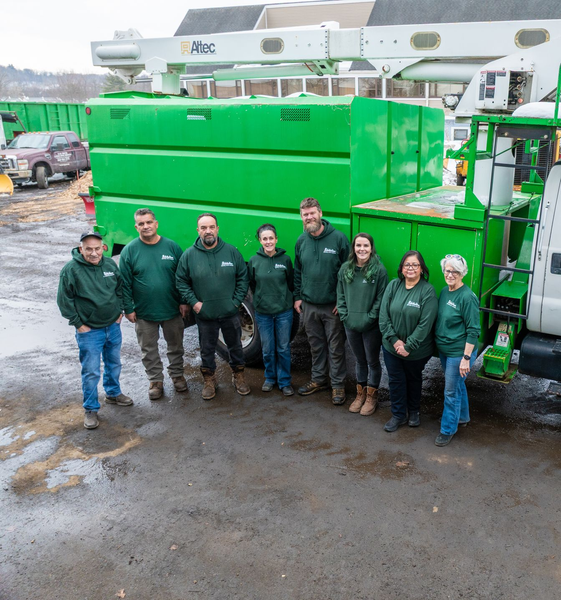A group of eight people in matching green hoodies stand in front of a large green work truck parked on a gravel lot.
