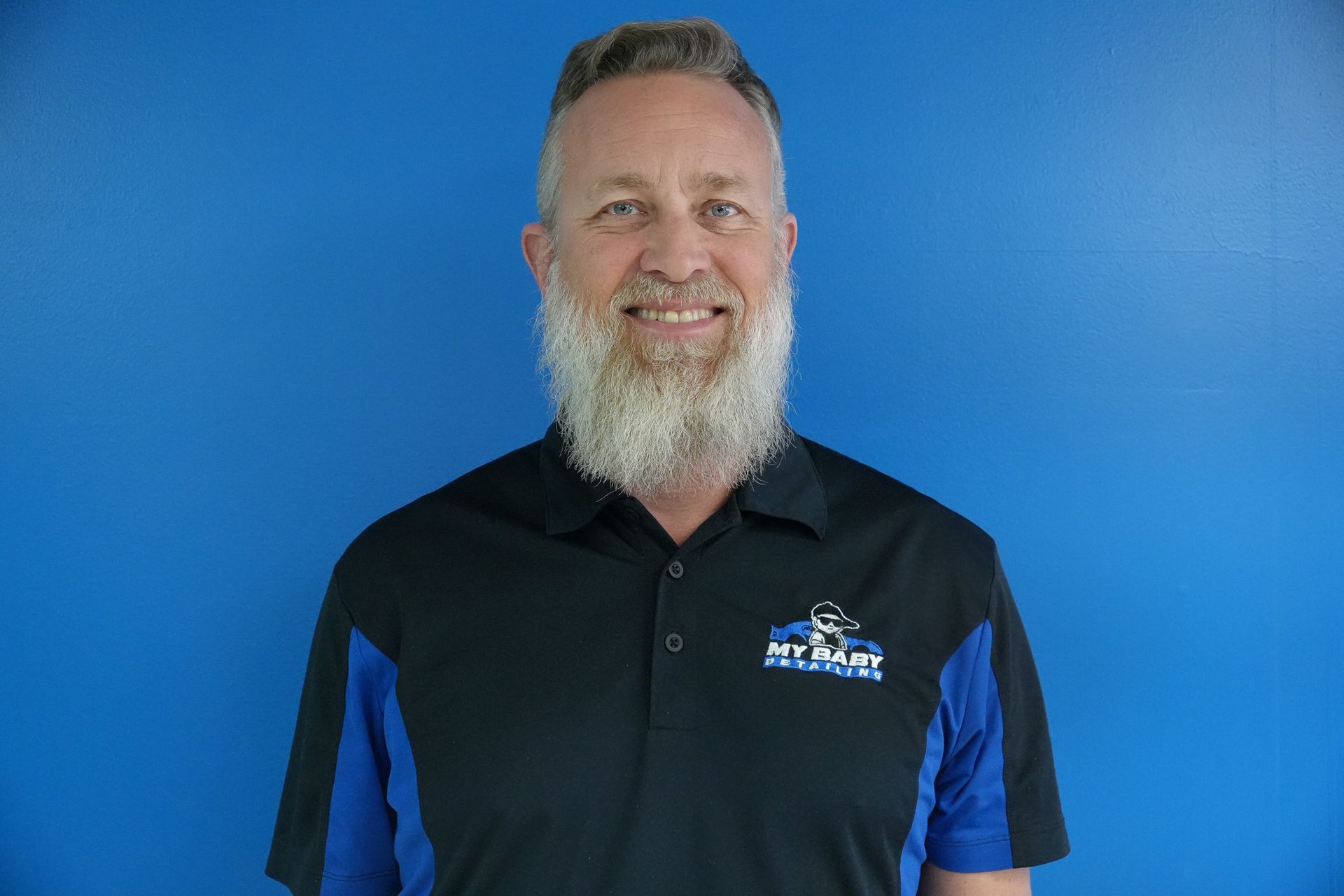 Man with a gray beard, wearing a black polo shirt with blue accents, smiles in front of a blue wall.