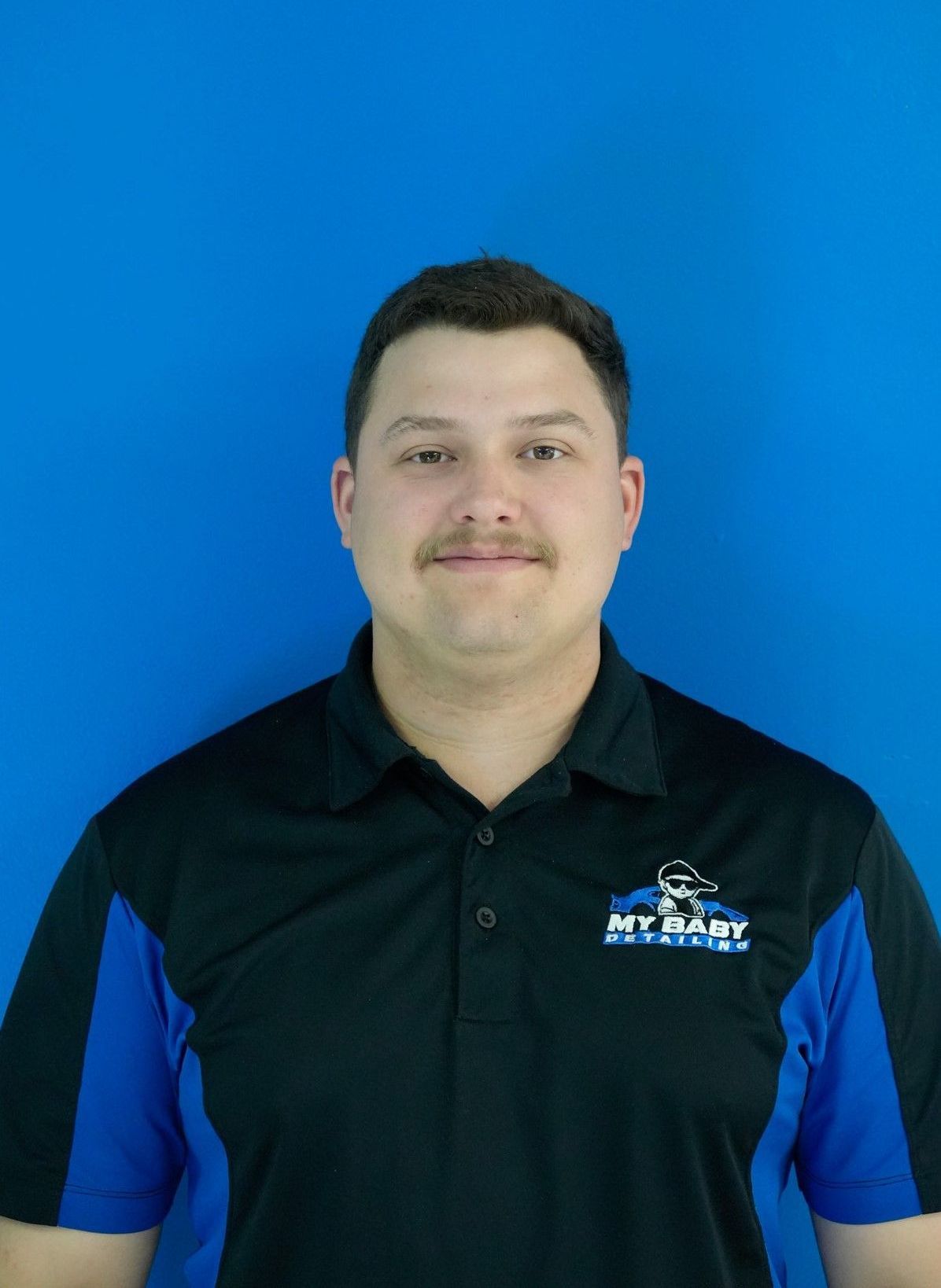 Man with dark hair and mustache wearing a black, blue, and white polo shirt against a blue background.