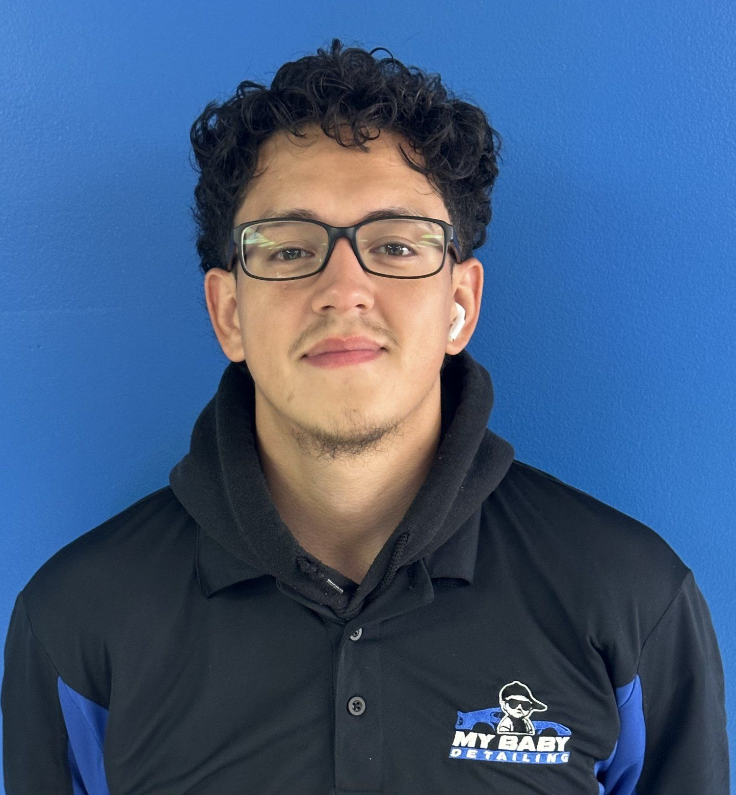 Man with curly hair and glasses smiles at the camera, blue wall background, wearing a black shirt with blue accents and logo.