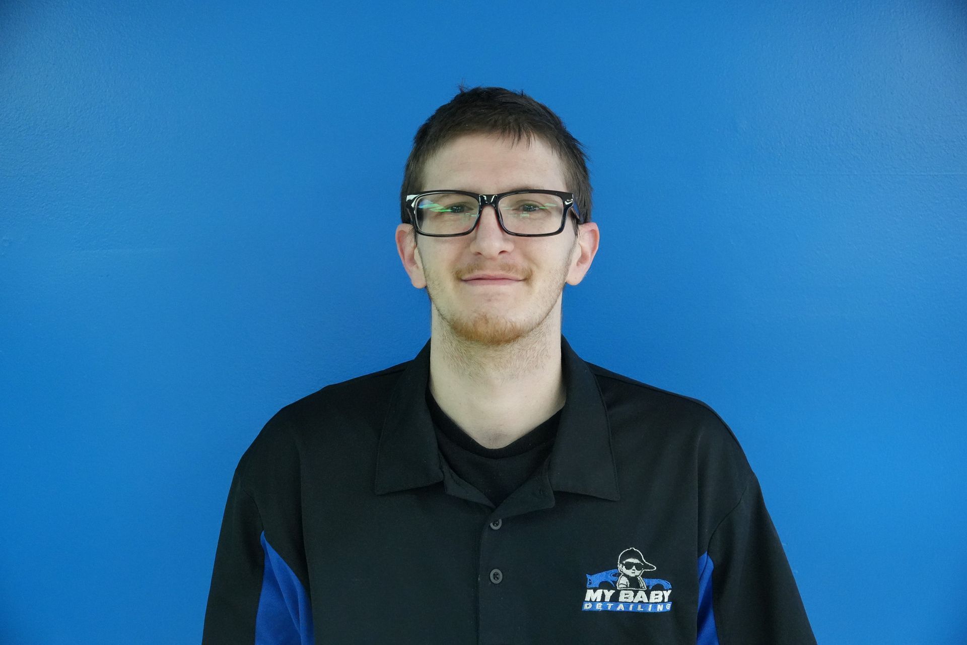 Man with glasses smiling in front of a blue backdrop, wearing a black polo shirt with a logo.