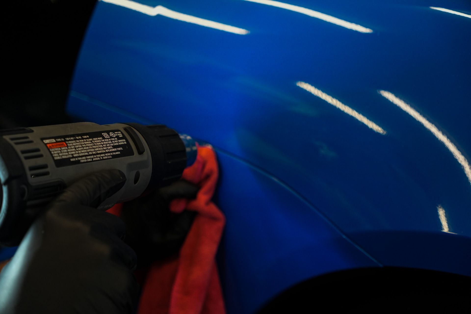 Gloved hand using a heat gun on blue car panel with a red cloth; indoors.