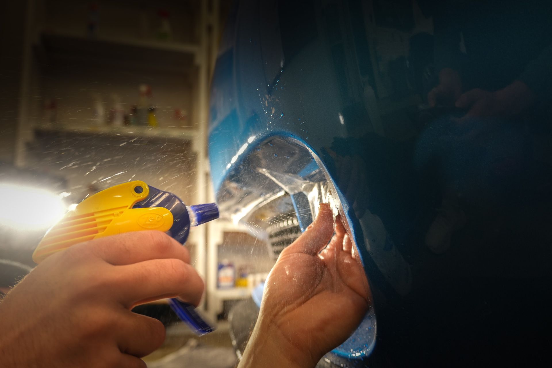Person sprays a clear protective film on a blue car's headlight with a spray bottle.