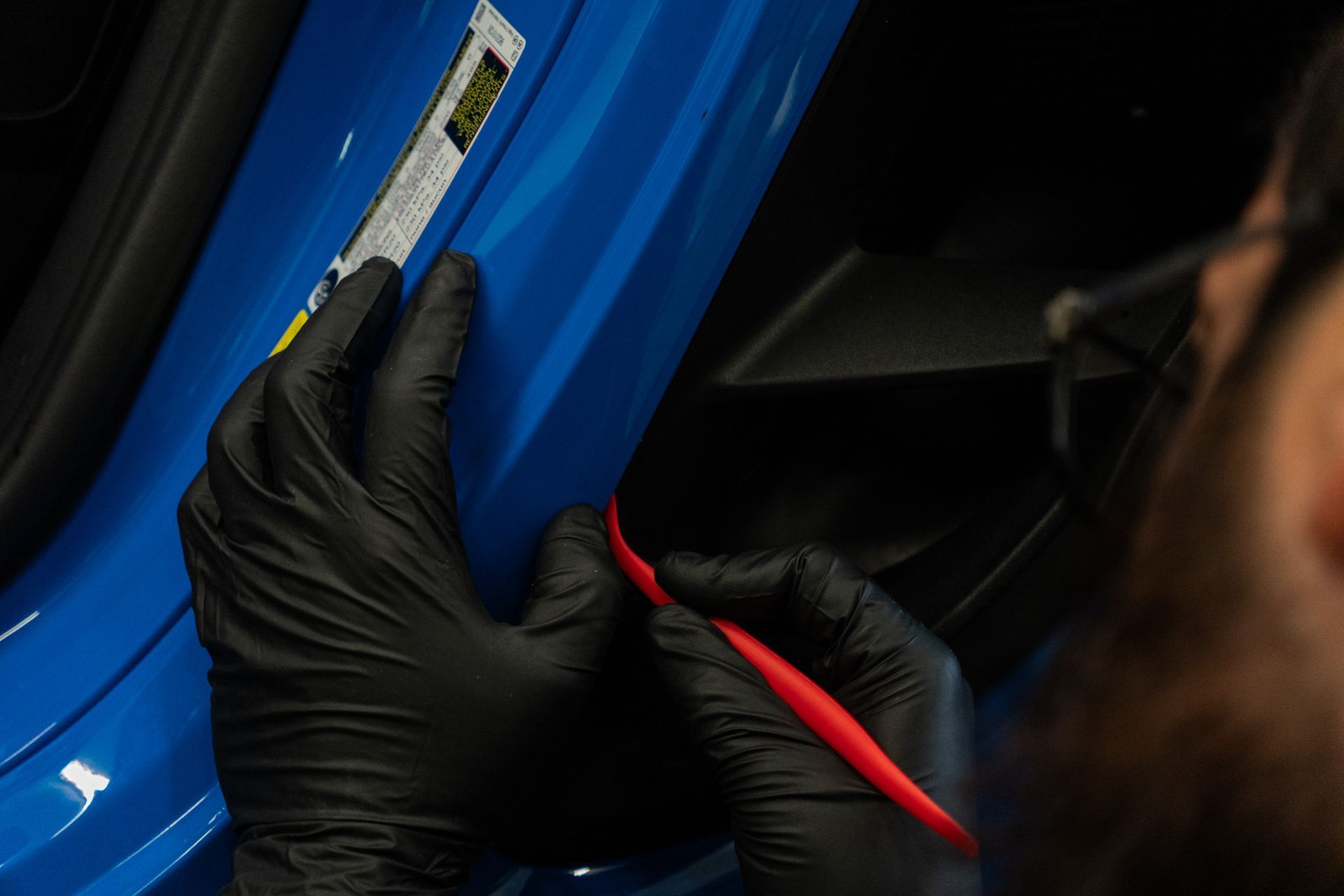 Person in black gloves using a red tool on a blue car door.