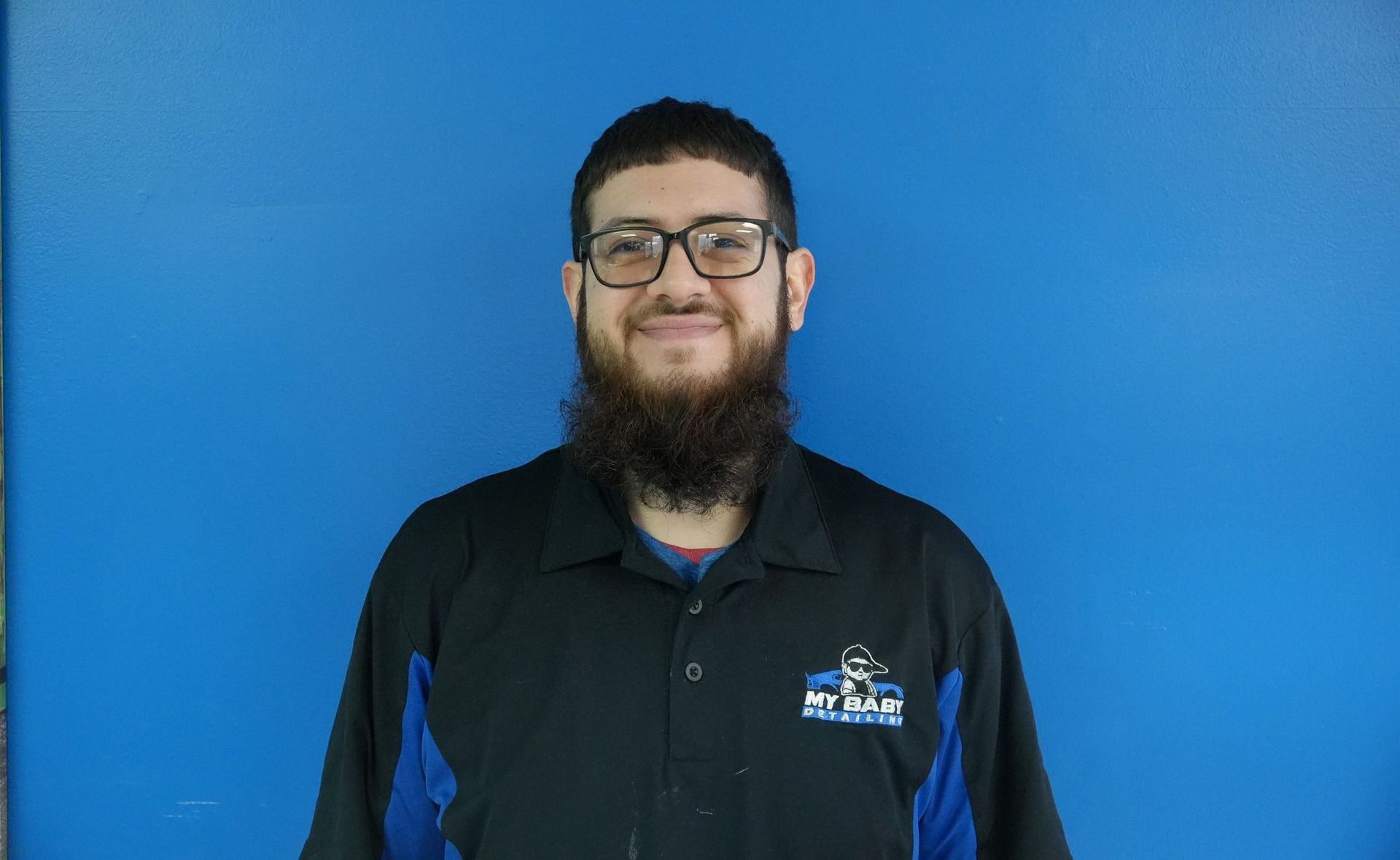 Man with glasses and beard in black and blue shirt smiles in front of a blue wall.