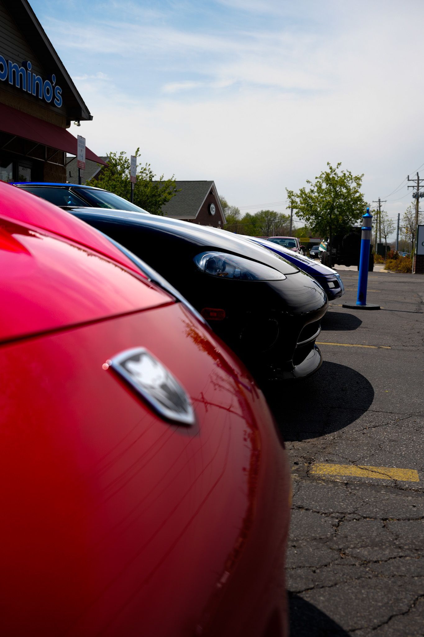 Red sports car in foreground, black and other cars parked in front of a Domino's on a sunny day.
