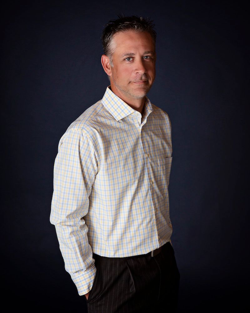 Man in a checkered shirt and dark pants, arms at his side. Dark blue background.