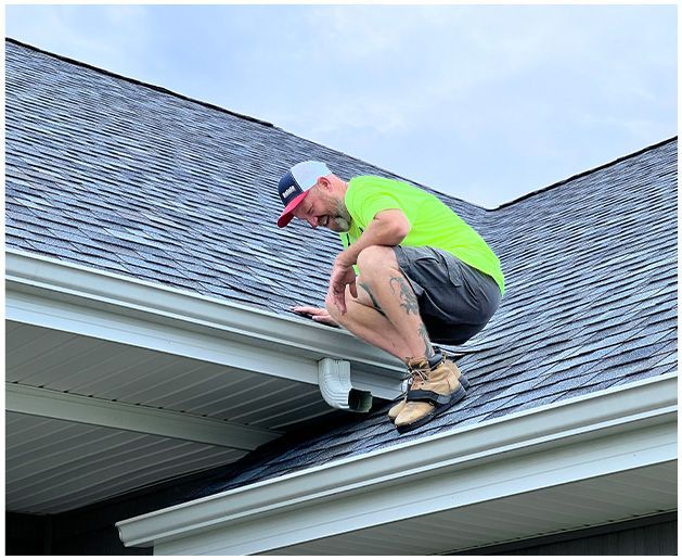 Man on a roof gutter, inspecting it; wearing neon green shirt, gray shorts, and a cap.