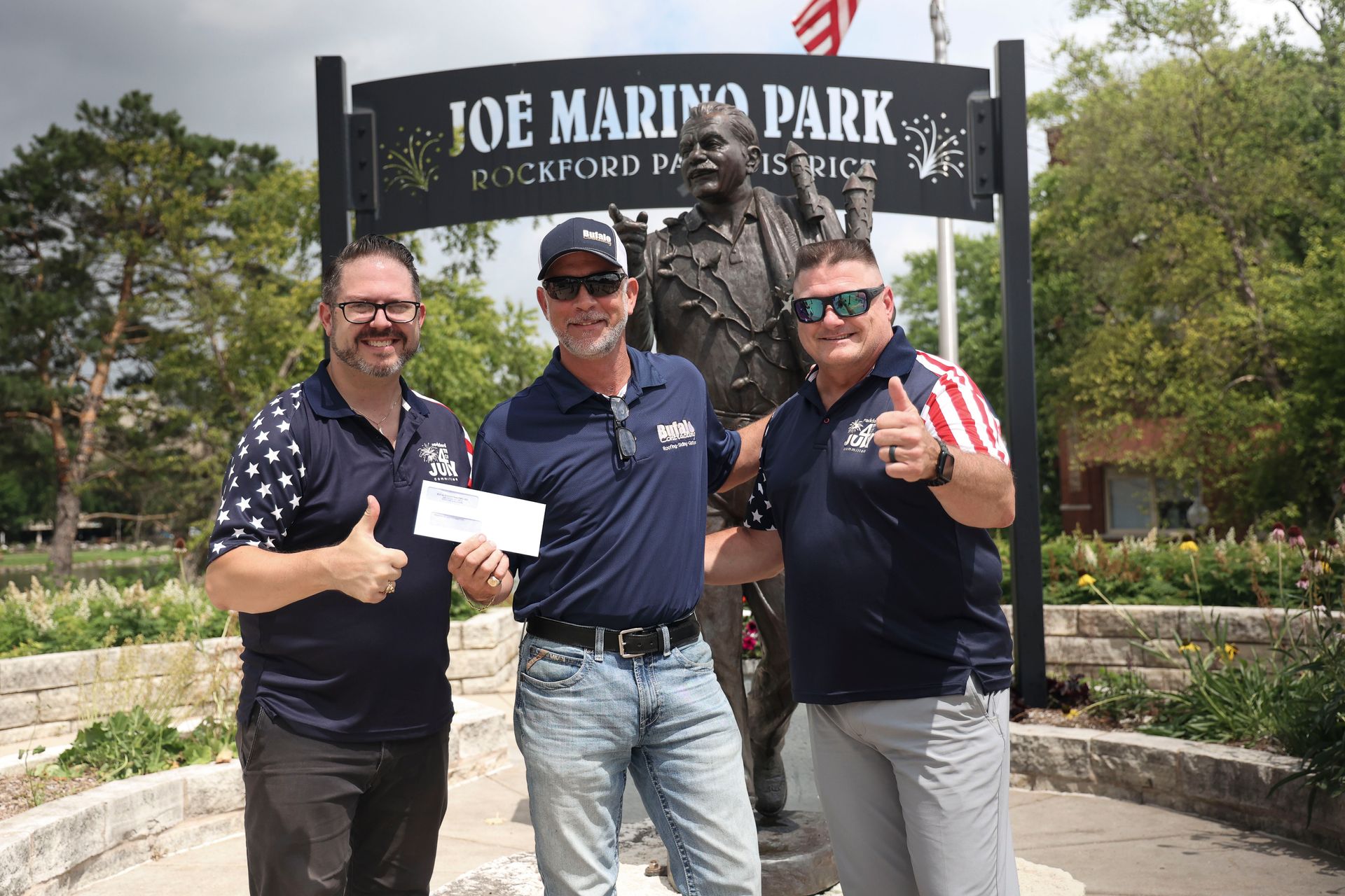 Three men at Joe Marino Park, posing near a statue. One holds a check, all give thumbs up.