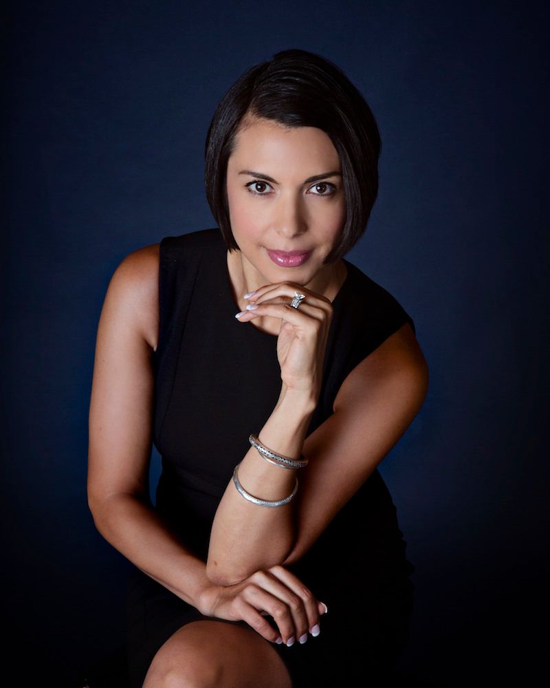 Woman in a black dress poses against a dark blue background, resting her chin on her hand, looking at the camera.