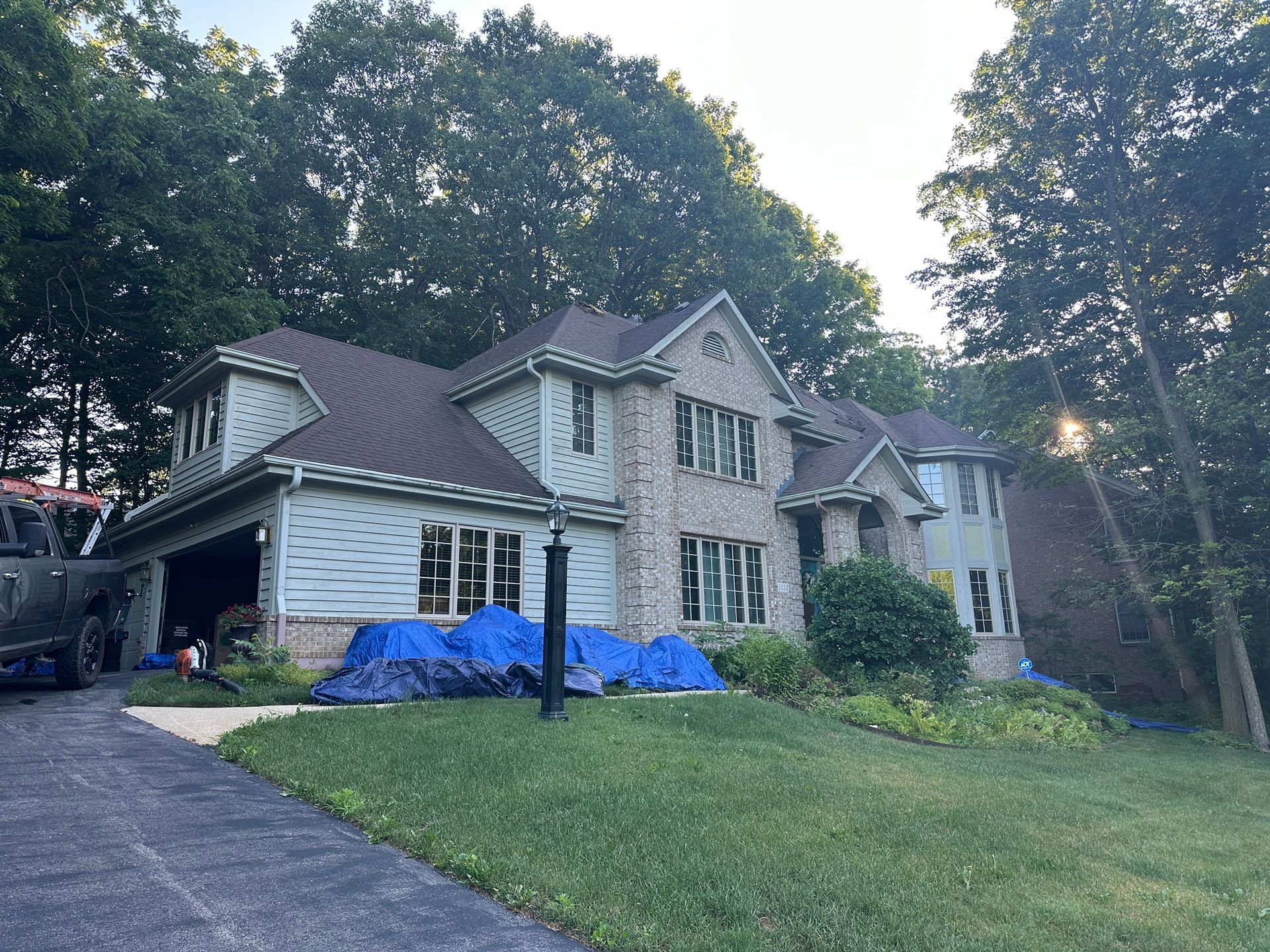 House with a dark roof, green siding, and brick facade. Tarps cover the landscaping; a truck is in the driveway.