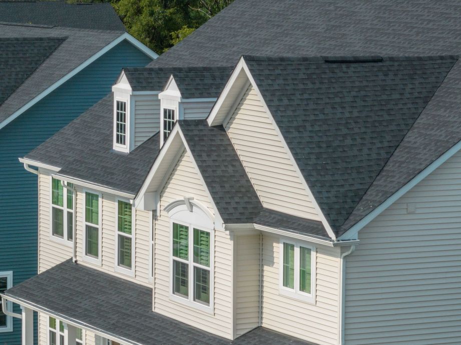 Beige house with dark gray roof, dormers, and white trim. Blue house in the background.