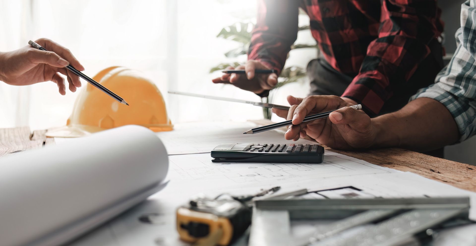 Three people collaborating over blueprints, with tools, a hard hat, and calculator on a table.