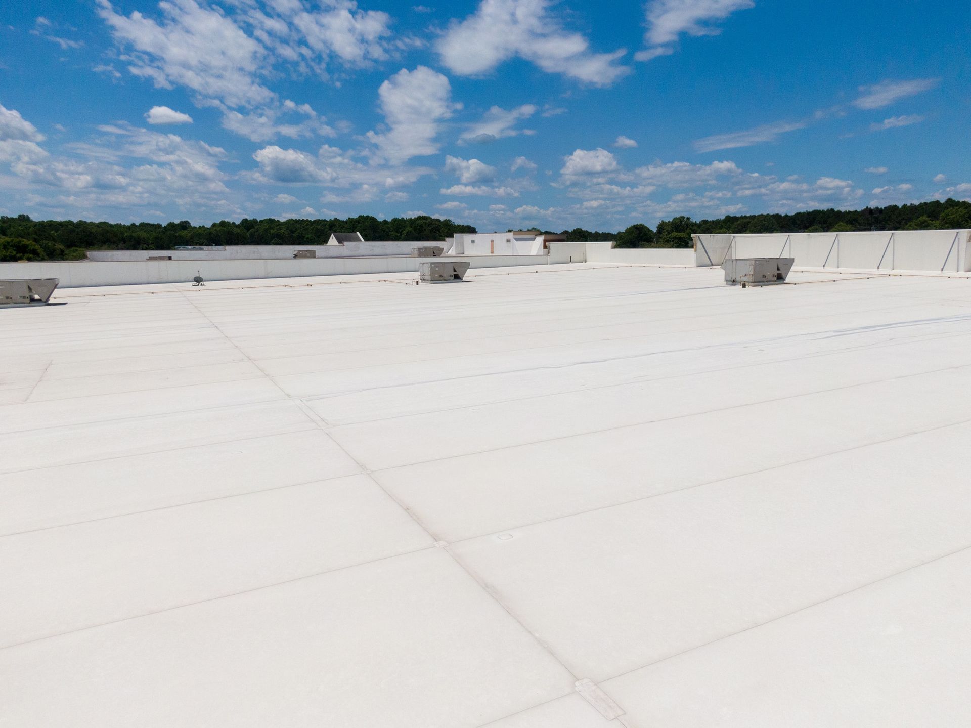 White commercial roof with air conditioning units, blue sky, and distant trees.