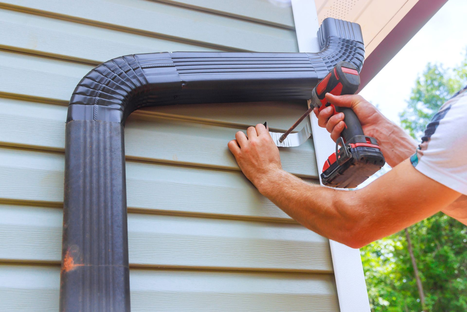 Person uses a power drill to attach a metal bracket to the siding near a black gutter.