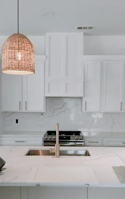 Modern white kitchen with a marble countertop, stainless steel sink, gold faucet, and a textured pendant light.