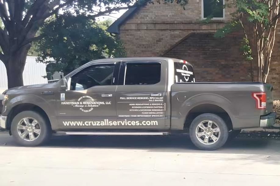 A dark gray pickup truck parked outdoors, featuring business logos and contact information for Cruz All Services on its side.