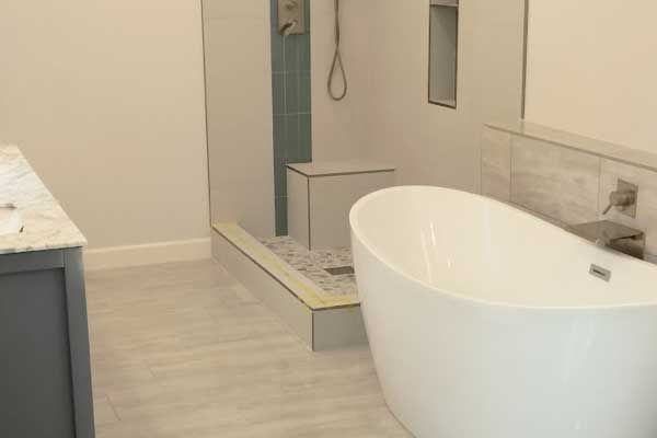A modern bathroom featuring a white freestanding soaking tub, a tiled shower with a bench, and gray cabinetry.