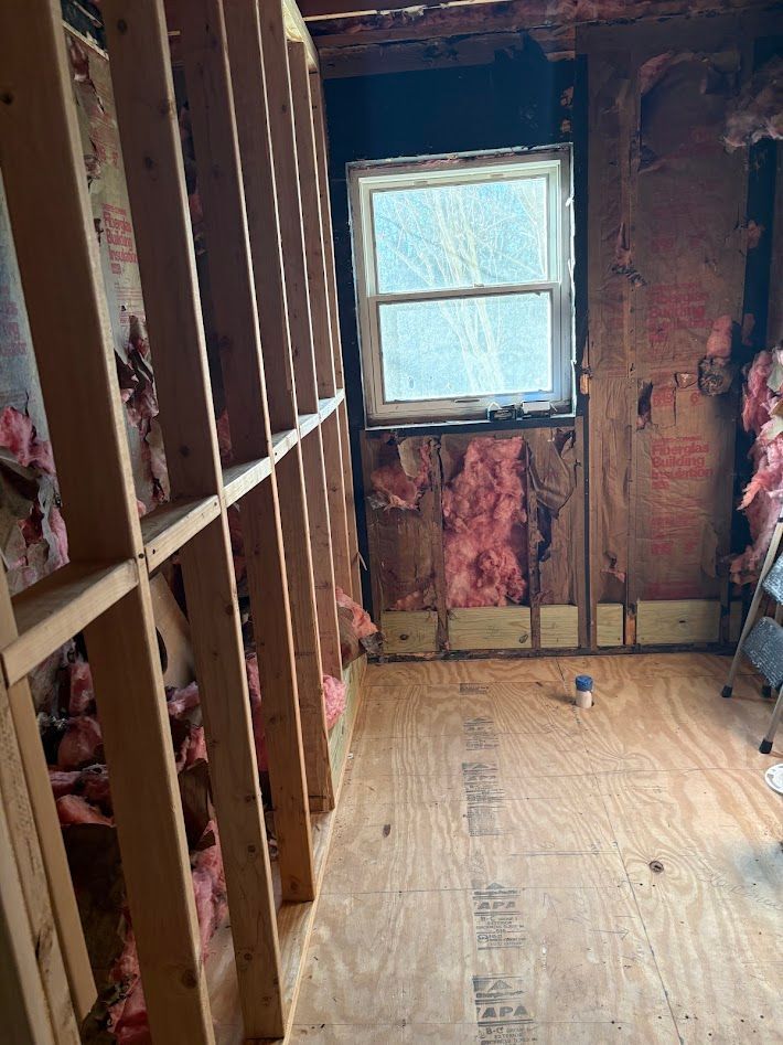 An interior view of a room under construction, showing wooden wall studs, pink fiberglass insulation, and a single window.