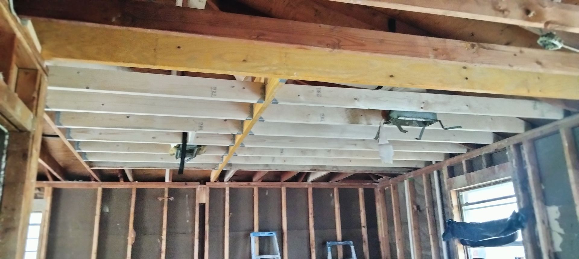 An interior view of a room under construction, showing exposed wooden ceiling joists, support beams, and wall framing.