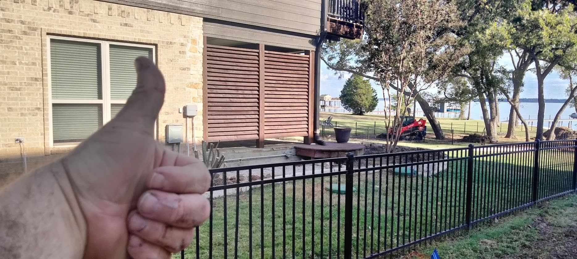 A person gives a thumbs-up in front of a brick house with a slatted wooden patio, metal fence, and yard by the water.