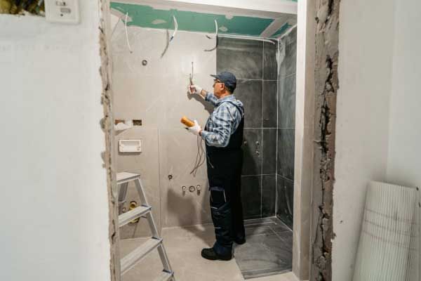 An electrician in workwear uses a multimeter to test wiring on a tiled bathroom wall during a renovation.