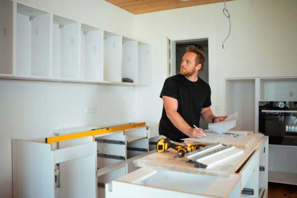 A person in a black shirt checks plans in a partially renovated kitchen with white cabinets and a yellow level.