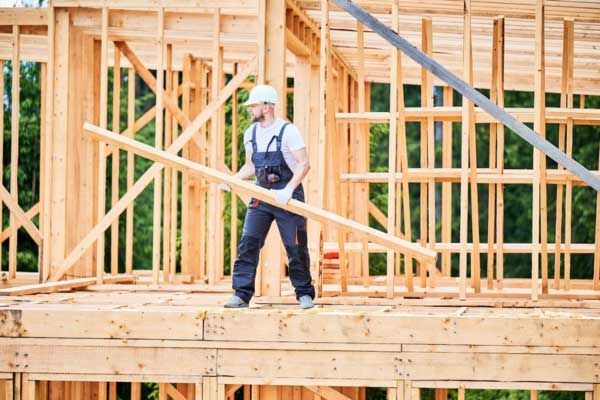 A construction worker in a white hard hat and workwear carries a wooden beam on the frame of a house under construction.